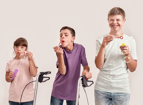 Three cheerful disabled children with Down syndrome and cerebral palsy smiling while blowing soap bubbles, standing together isolated over white background. Lifestyle of special children concept
