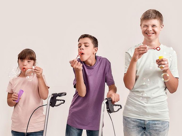 Three cheerful disabled children with Down syndrome and cerebral palsy smiling while blowing soap bubbles, standing together isolated over white background. Lifestyle of special children concept