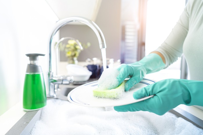 Close up of woman hands  washing a plate with a cleaning sponge in a kitchen sink. A faucet with flowing water and a dish washing soap are visible. The sink is full of water and soap sud. High resolution 42Mp indoors digital capture taken with SONY A7rII and Zeiss Batis 25mm F2.0 lens