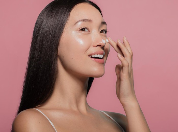 1142851446 Close up of a youthful female model applying moisturizer to her face. Young korean woman putting moisturizer cream on her pretty face against pink background.