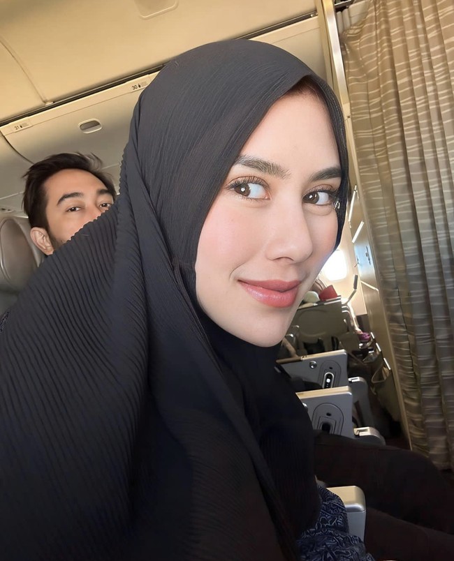 Pose Syahnaz pakai hijab instan bahan crinkle atau plisket warna hitam saat di pesawat. Terlihat Syahnaz duduk di samping sang suami, Jeje ‘Govinda’. Foto: Instagram @syahnaz 