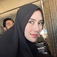 Pose Syahnaz pakai hijab instan bahan crinkle atau plisket warna hitam saat di pesawat. Terlihat Syahnaz duduk di samping sang suami, Jeje ‘Govinda’. Foto: Instagram @syahnaz 