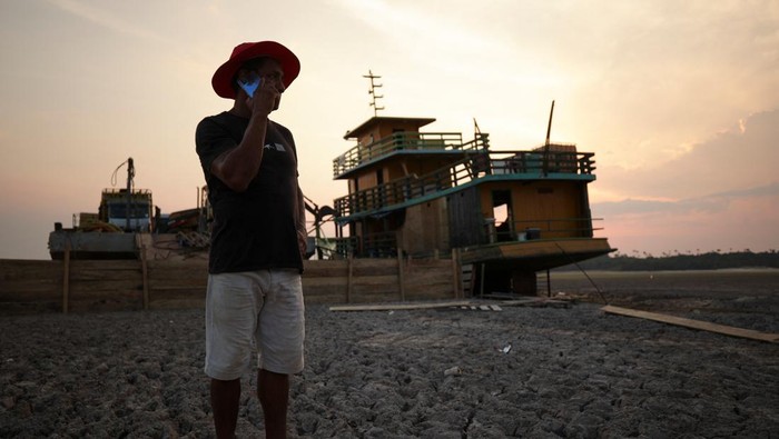 Penampakan Kapal Tongkang Terdampar di Sungai Amazon yang Mengering A tug boat and a barge carrying three trucks, 2,000 empty cooking gas cylinders and a backhoe, lie stranded on a sand bank of a diminished Rio Negro river after running aground last month, as the region is hit by a severe drought, in Cacau Pirera, Brazil October 10, 2023. REUTERS/Bruno Kelly