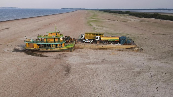 Penampakan Kapal Tongkang Terdampar di Sungai Amazon yang Mengering A tug boat and a barge carrying three trucks, 2,000 empty cooking gas cylinders and a backhoe, lie stranded on a sand bank of a diminished Rio Negro river after running aground last month, as the region is hit by a severe drought, in Cacau Pirera, Brazil October 10, 2023. REUTERS/Bruno Kelly