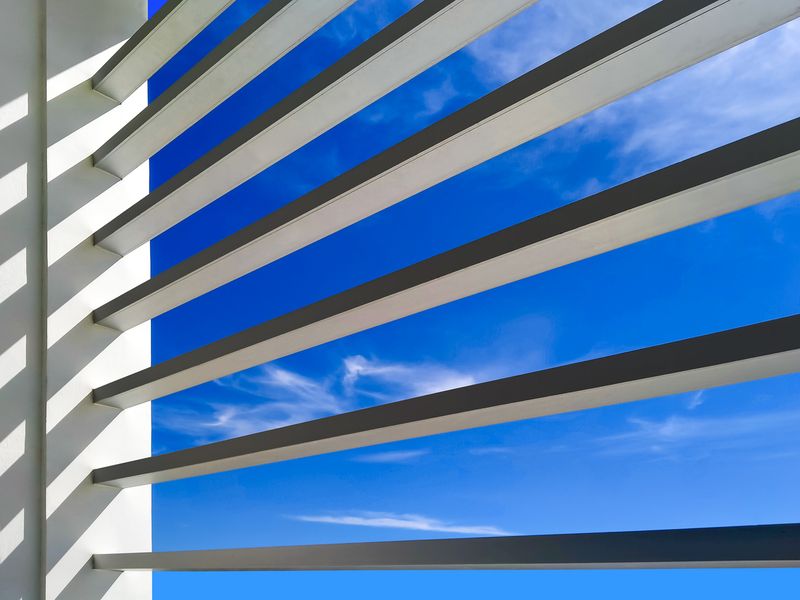 Sunlight on sunshade battens surface of office building against blue sky background, low angle view from inside of building