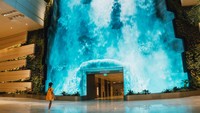 Di aula keberangkatan T2, terdapat The Wonderfall, kanvas digital setinggi 14-meter yang terletak di tengah taman vertikal, menampilkan keindahan alam dengan air terjun megah yang mengalir di atas batu-batu megalitik. Sungguh menakjubkan! (dok. Changi Airport Group)