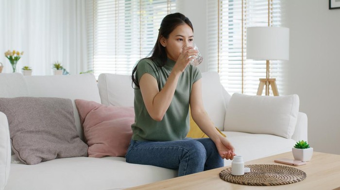 Sick Asian woman eating pills with a glass of water in hand near window in her house.
