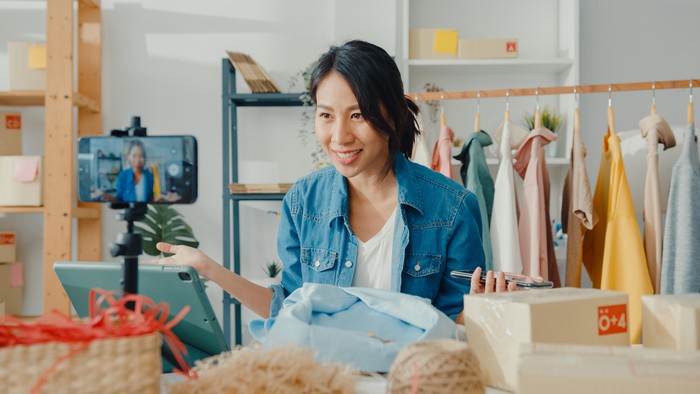 Young Asia lady fashion designer using mobile phone receiving purchase order and showing clothes recording video live streaming online at shop. Small business owner, online market delivery concept.