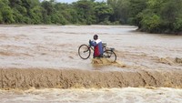 Nampak seorang warga menyeberangi sungai yang meluap dengan membawa sepeda.  