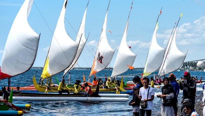 Puluhan perahu dengan layar berkibar mengarungi Selat Bali. Perahu layar tradisional tersebut berpacu dengan ombak laut Bali.