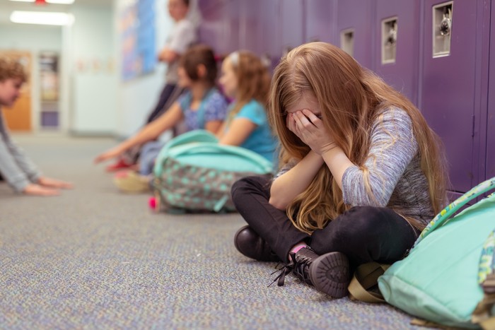 A sad elementary-age girl sits by her locker with her face in her hands. She is crying and classmates in the background are talking and playing with each other.
