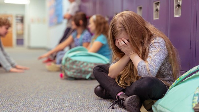 A sad elementary-age girl sits by her locker with her face in her hands. She is crying and classmates in the background are talking and playing with each other.