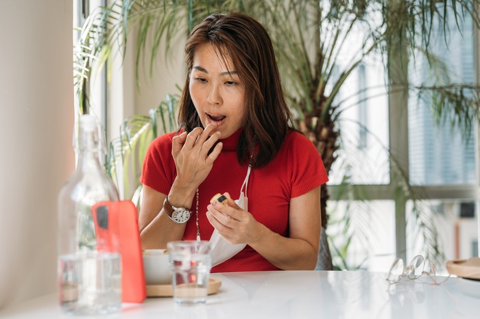 Ilustrasi bibir An Asian Chinese woman putting on lip balm using smartphone as a mirror at office.