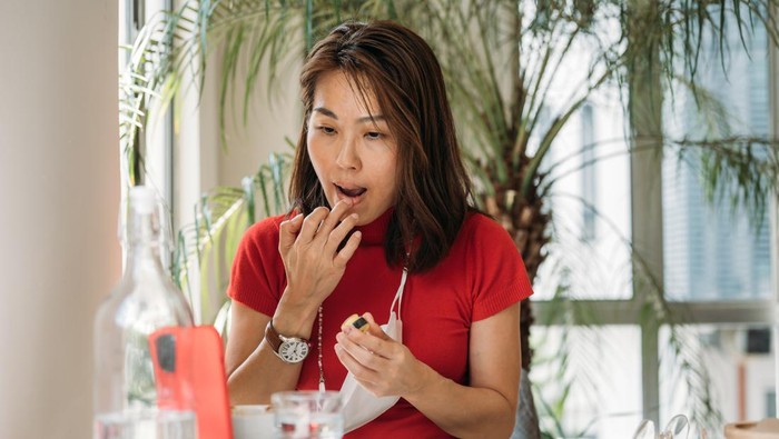 An Asian Chinese woman putting on lip balm using smartphone as a mirror at office.