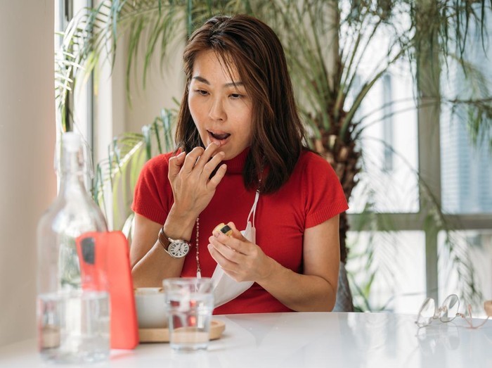 An Asian Chinese woman putting on lip balm using smartphone as a mirror at office.