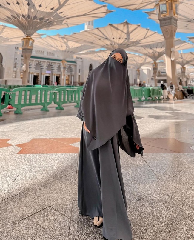Ria Ricis mengunggah ke Instagram foto saat sedang berada di halaman Masjid Nabawi, Madinah. Ricis mengenakan french khimar, cadar dan abaya crinkle warna abu-abu. Ricis mulai berada di Tanah Suci sejak 31 Desember 2023. Foto: Dok. Instagram @riaricis1795.