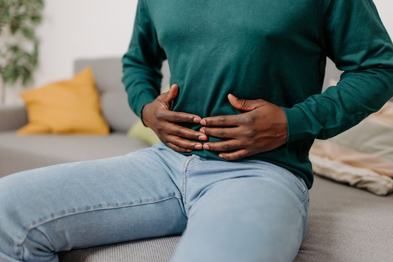 This image portrays a young African American man sitting on a sofa in his living room, holding his stomach due to digestive problems