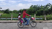 Seorang ibu sedang memboceng anaknya melintasi jembatan dengan latar belakang Sungai Prasung penuh dengan eceng gondok.