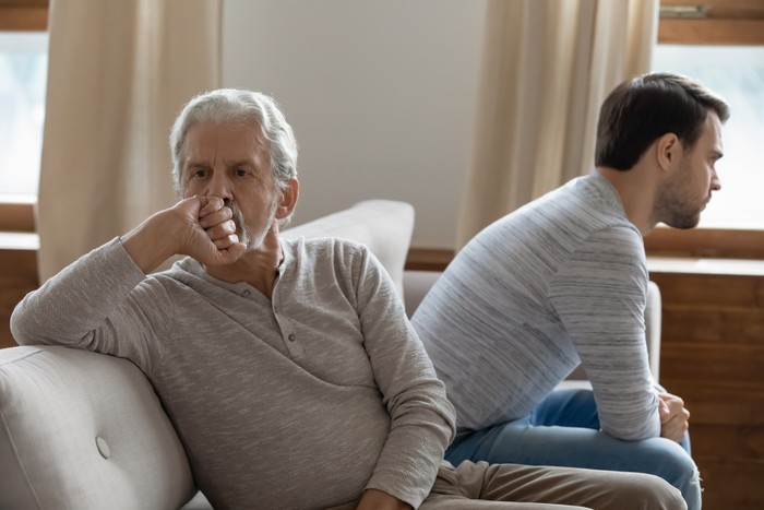 Unhappy mature father and adult son not taking after quarrel, sitting on couch at home separately back to back, upset older grandfather and grandson ignoring each other, two generations conflict