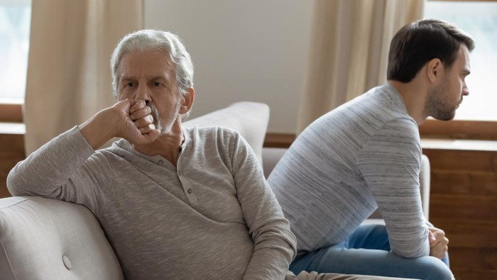 Unhappy mature father and adult son not taking after quarrel, sitting on couch at home separately back to back, upset older grandfather and grandson ignoring each other, two generations conflict