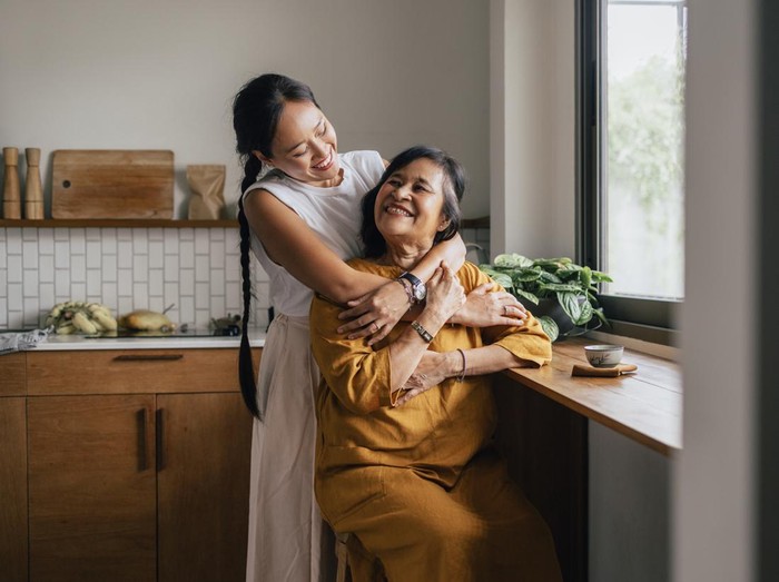 1459436747 A smiling Asian woman enjoying drinking tea while her loving daughter is embracing her.
