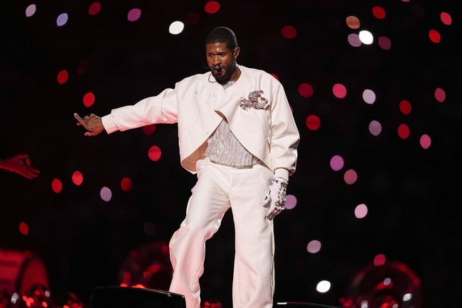 Usher mengawali penampilannya dengan kombinasi busana serba putih yang dirancang khusus oleh Dolce Gabbana. Busana tersebut terdiri dari crop putih yang dihiasi detail bebatuan, celana senada, kemudian dibungkus jaket stylish dan coat dramatis. (Foto: AP Photo/Brynn Anderson)