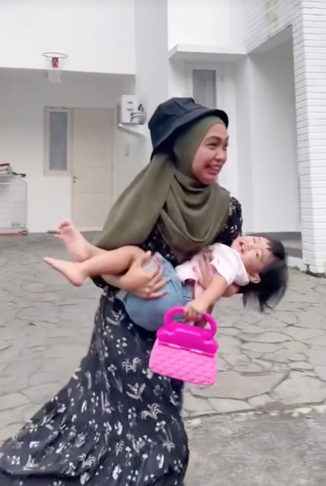 Tak ketinggalan Ria Ricis juga ikutan membuat video parodi aksi Mayor Teddy. Kali ini yang digendong Ricis ialah anaknya sendiri, Moana. Moana yang sedang berdiri membawa tas pink itu tertawa usai tiba-tiba digendong oleh sang bunda. Foto: TikTok