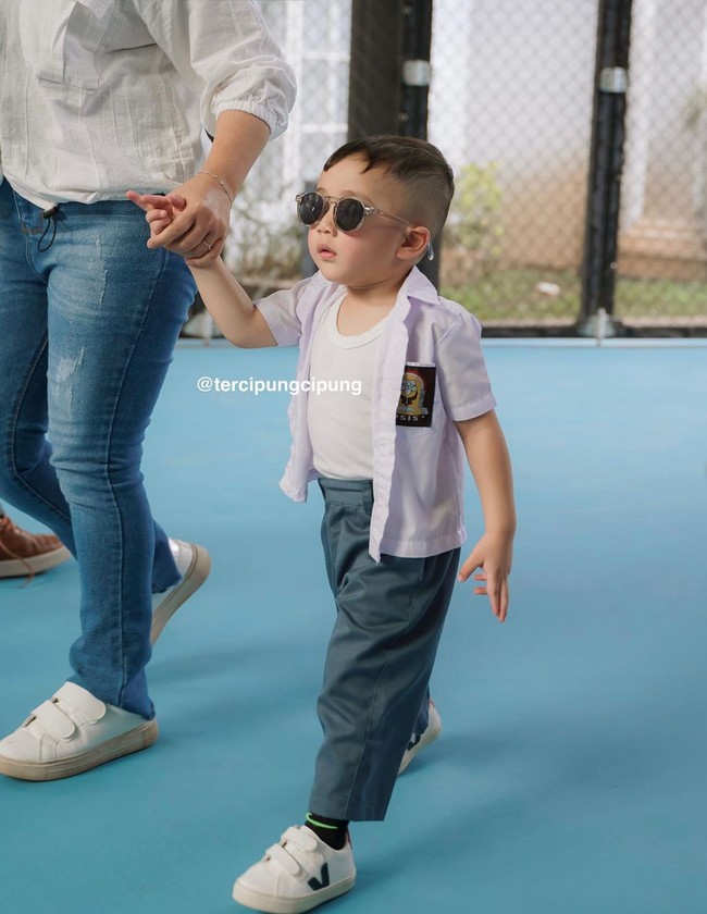 Rayyanza tampil mengenakan seragam SMA putih abu-abu yang dipadukan dengan sneakers putih dan kaca mata hitam. Kemeja yang dikenakannya itu tampak sengaja dibuka dan memperlihatkan kaos dalamnya. Tampil bergaya bak ketua geng, Rayyanza sukses curi atensi. Foto: Instagram/@raffinagita1717