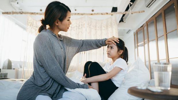 Sick little daughter lying in bed. A loving Asian mother checking temperature on her forehead. A glass of water and medicine on the side table
