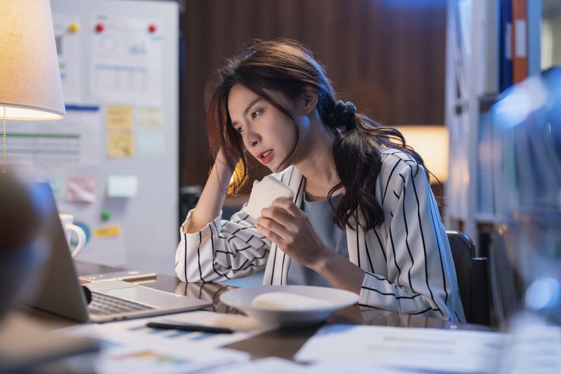 Young asian female adult woman having a sandwich while super busy working from home late at night she is working hard to finish her project tonight