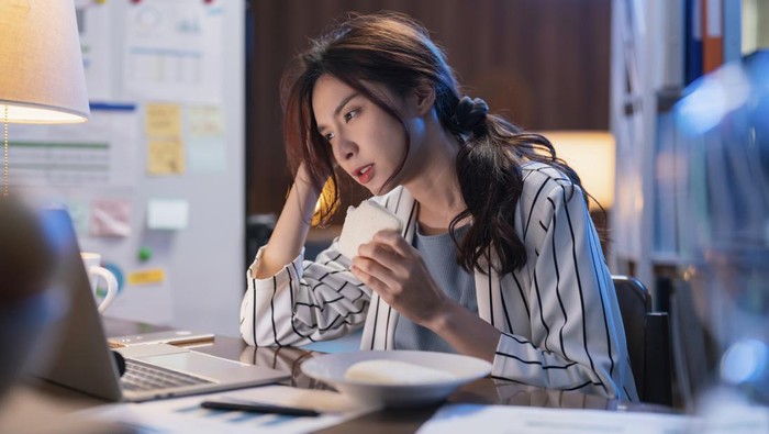 Young asian female adult woman having a sandwich while super busy working from home late at night she is working hard to finish her project tonight