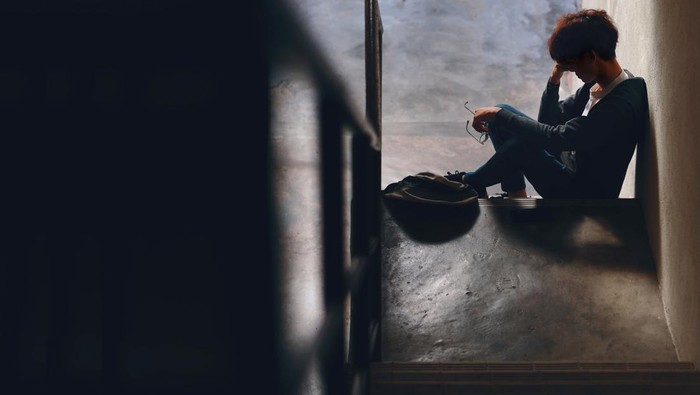 An upset Asian teenage boy student, dressed in casual outfit, sits on a stairway reading on his smartphone with a backpack beside him, while dealing with the issue of cyberbullying.