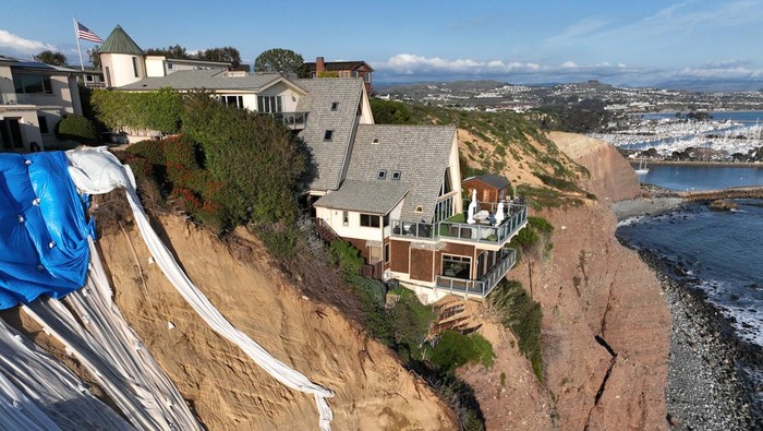 Dana Point, CA - February 27: An aerial view of a double A-frame cliff-side mansion, between two other mansions affected by a landslide and protective tarps to protect from further slides from upcoming storms in Dana Point Tuesday, Feb. 27, 2024. The three homes affected by recent rain storms are being monitored but officials say they don't appear to be in imminent danger. (Allen J. Schaben / Los Angeles Times via Getty Images)