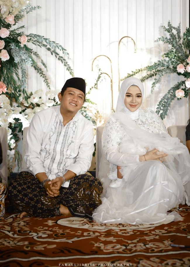 Wanita asal Madiun ini memilih gaun putih dengan aksen kristal, bordir dan payet yang dipadankan jilbab putih. Senada dengan sang istri, Denny Caknan memakai baju koko berhias bordiran yang dipadankan kain batik dan peci. Foto: Instagram/@denny_caknan @bellabonita_r.a