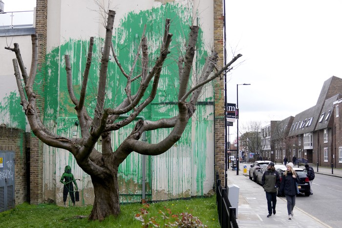 A woman takes a picture on her phone of a new Banksy painting on a wall in London, Monday, March 18, 2024. A new Banksy mural drew crowds to a London street on Monday, even before the elusive graffiti artist confirmed that the work was his. The artwork in the Finsbury Park neighborhood covers the wall of a four-story building and shows a small figure holding a pressure hose beside a real tree. Green paint has been sprayed across the wall, replicating the absent leaves of the tree, which has been severely cropped. Banksy claimed the work by posting before and after photos of the location on his official Instagram account.(AP Photo/Alastair Grant)