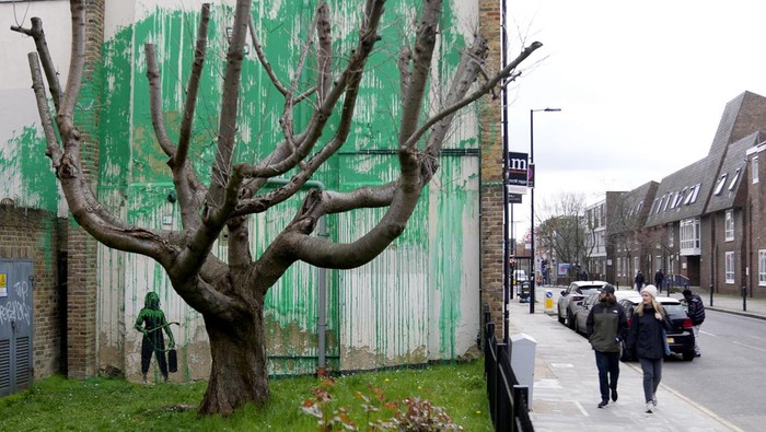 A woman takes a picture on her phone of a new Banksy painting on a wall in London, Monday, March 18, 2024. A new Banksy mural drew crowds to a London street on Monday, even before the elusive graffiti artist confirmed that the work was his. The artwork in the Finsbury Park neighborhood covers the wall of a four-story building and shows a small figure holding a pressure hose beside a real tree. Green paint has been sprayed across the wall, replicating the absent leaves of the tree, which has been severely cropped. Banksy claimed the work by posting before and after photos of the location on his official Instagram account.(AP Photo/Alastair Grant)