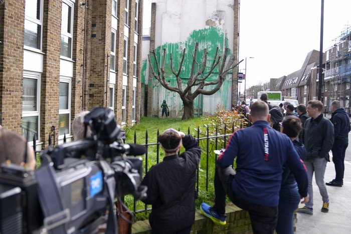 A woman takes a picture on her phone of a new Banksy painting on a wall in London, Monday, March 18, 2024. A new Banksy mural drew crowds to a London street on Monday, even before the elusive graffiti artist confirmed that the work was his. The artwork in the Finsbury Park neighborhood covers the wall of a four-story building and shows a small figure holding a pressure hose beside a real tree. Green paint has been sprayed across the wall, replicating the absent leaves of the tree, which has been severely cropped. Banksy claimed the work by posting before and after photos of the location on his official Instagram account.(AP Photo/Alastair Grant)