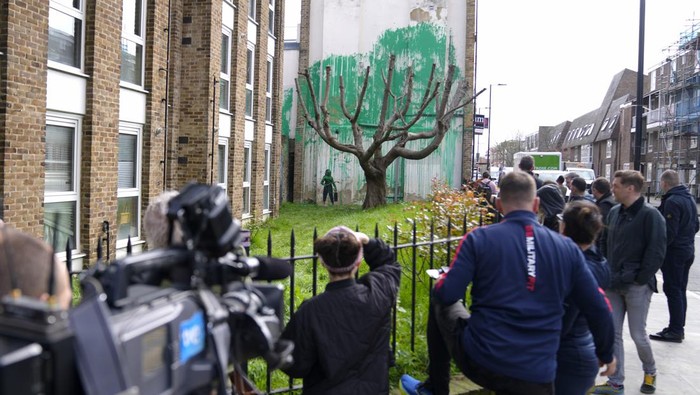A woman takes a picture on her phone of a new Banksy painting on a wall in London, Monday, March 18, 2024. A new Banksy mural drew crowds to a London street on Monday, even before the elusive graffiti artist confirmed that the work was his. The artwork in the Finsbury Park neighborhood covers the wall of a four-story building and shows a small figure holding a pressure hose beside a real tree. Green paint has been sprayed across the wall, replicating the absent leaves of the tree, which has been severely cropped. Banksy claimed the work by posting before and after photos of the location on his official Instagram account.(AP Photo/Alastair Grant)