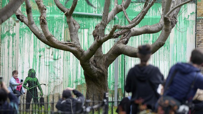 A woman takes a picture on her phone of a new Banksy painting on a wall in London, Monday, March 18, 2024. A new Banksy mural drew crowds to a London street on Monday, even before the elusive graffiti artist confirmed that the work was his. The artwork in the Finsbury Park neighborhood covers the wall of a four-story building and shows a small figure holding a pressure hose beside a real tree. Green paint has been sprayed across the wall, replicating the absent leaves of the tree, which has been severely cropped. Banksy claimed the work by posting before and after photos of the location on his official Instagram account.(AP Photo/Alastair Grant)