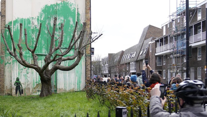 A woman takes a picture on her phone of a new Banksy painting on a wall in London, Monday, March 18, 2024. A new Banksy mural drew crowds to a London street on Monday, even before the elusive graffiti artist confirmed that the work was his. The artwork in the Finsbury Park neighborhood covers the wall of a four-story building and shows a small figure holding a pressure hose beside a real tree. Green paint has been sprayed across the wall, replicating the absent leaves of the tree, which has been severely cropped. Banksy claimed the work by posting before and after photos of the location on his official Instagram account.(AP Photo/Alastair Grant)