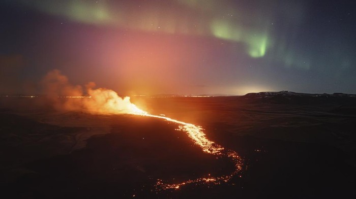 A view of the lava flowing from the volcano backdropped by the Northern Lights, near the town of Grindavik, Iceland, early Saturday, March 23, 2024.  The volcano in southwestern Iceland that erupted three times in December, January and February, sending lava towards a nearby community, keeps erupting. (AP Photo/Marco di Marco)