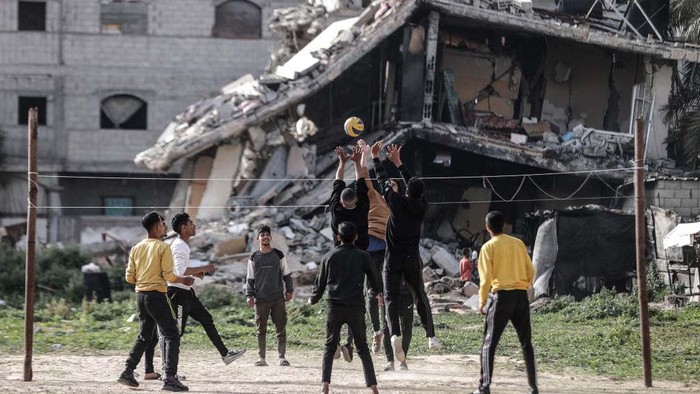 Warga Gaza Bermain Voli di Samping Reruntuhan Bangunan DEIR AL-BALAH, GAZA - MARCH 27: Palestinian youths, who take refuge in Nuseirat camp from ongoing Israeli attacks, pass the time by playing volleyball near a partially collapsed building while waiting for iftar time on the Holy month of Ramadan in Deir al-Balah, Gaza on March 27, 2024. (Photo by Ali Jadallah/Anadolu via Getty Images)