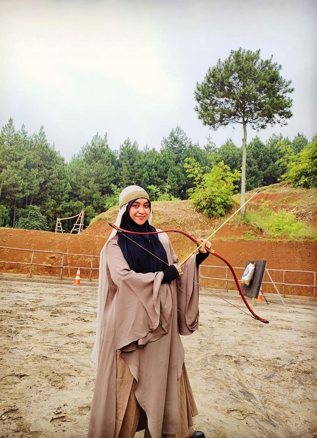 Selain berdakwah, Haneen juga menyalurkan hobi memanah. Haneen berpose dengan busur dan anak panah. Haneen memakai dress warna cokelat dipadu dengan hijab syari hitam. Foto: Dok. Instagram @haneenakira.