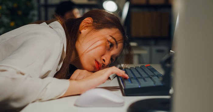 Closeup of Asian woman appears deeply fatigued and stressed while working on laptop in office night, highlighting issues of overwork and employee burnout. ESG sustainable business office concept.