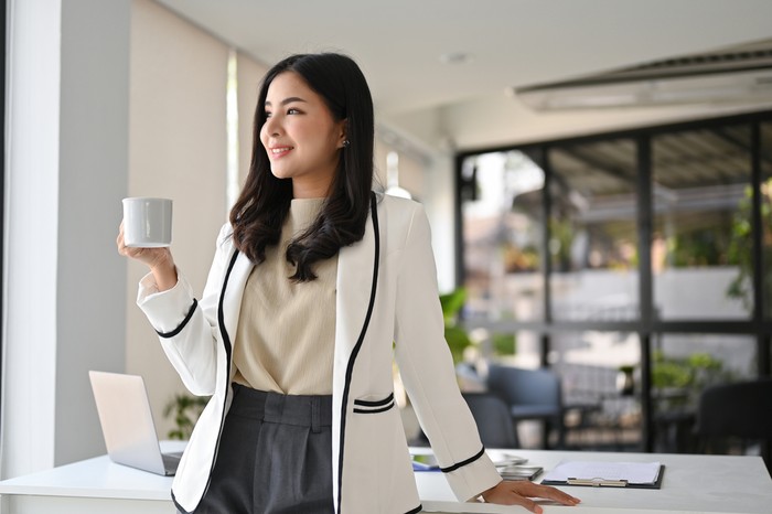 ilustrasi empowering women wanita karier Attractive and successful millennial Asian businesswoman in white suit stands in her office looking out the window and daydreaming about her career success while sipping coffee.