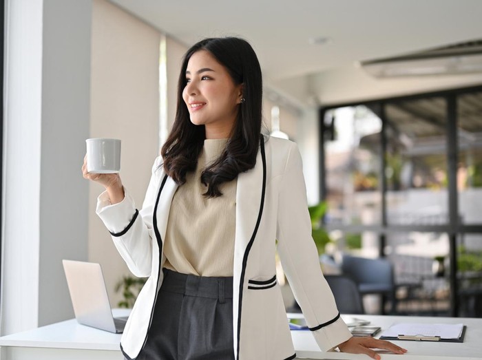 Attractive and successful millennial Asian businesswoman in white suit stands in her office looking out the window and daydreaming about her career success while sipping coffee.