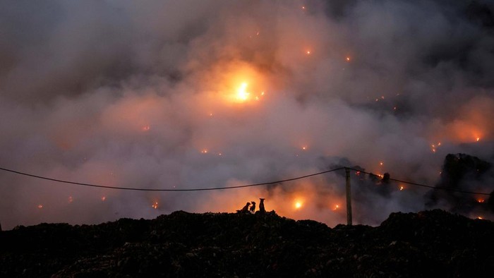 People sit at a tea stall inside a fish market as fire billows from burning garbage at the Ghazipur landfill site, in New Delhi, India, April 22, 2024. REUTERS/Adnan Abidi