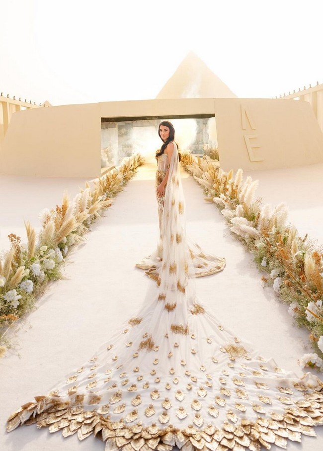 Meskipun dekorasi pernikahan minimalis, sang mempelai wanita, Erika Hammond, tampil megah dalam balutan gaun bergaya ekstravaganza. Wanita 33 tahun ini mengenakan gaun pengantin rancangan Rahul Mishra. Gaun transparan berbahan tulle dihiasi ornamen emas, serta dilengkapi selendang yang memanjang dengan motif dedaunan menambah kesan dramatis. Foto: Instagram/@erikaannhammond @bottega53