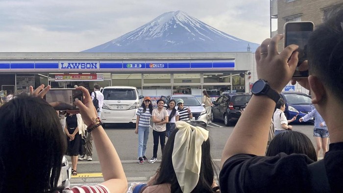 Visitors take a photo in front of a convenient store at Fujikawaguchiko town, Yamanashi prefecture, Japan, with a backdrop of Mr. Fuji on April 28, 2024. The town of Fujikawaguchiko, known for a number of popular photo spots for Japans trademark of Mt. Fuji, on Tuesday, April 30, 2024 began to set up a huge black screen on a stretch of sidewalk to block view of the mountain in a neighborhood hit by a latest case of overtourism in Japan. (Kyodo News via AP)