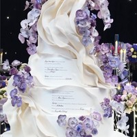 Kue pengantin pun diberi sentuhan personal. LeNovelle Cake mengkreasikan wedding cake putih berhias bunga anggrek dengan penggalan lirik lagu duet mereka. Foto: Instagram @thebridestory @johan.k.lotus